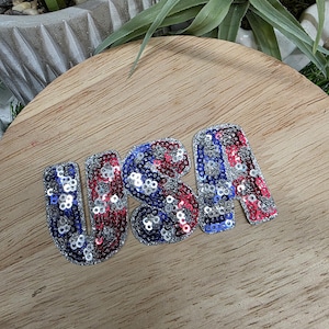 Pu&ograve; includere: Un emblema "USA" con paillettes rosse, bianche e blu, che ricorda la bandiera americana. Le lettere sono decorate con paillettes lucenti e sono poste su una superficie di legno. Si tratta di un oggetto decorativo patriottico.