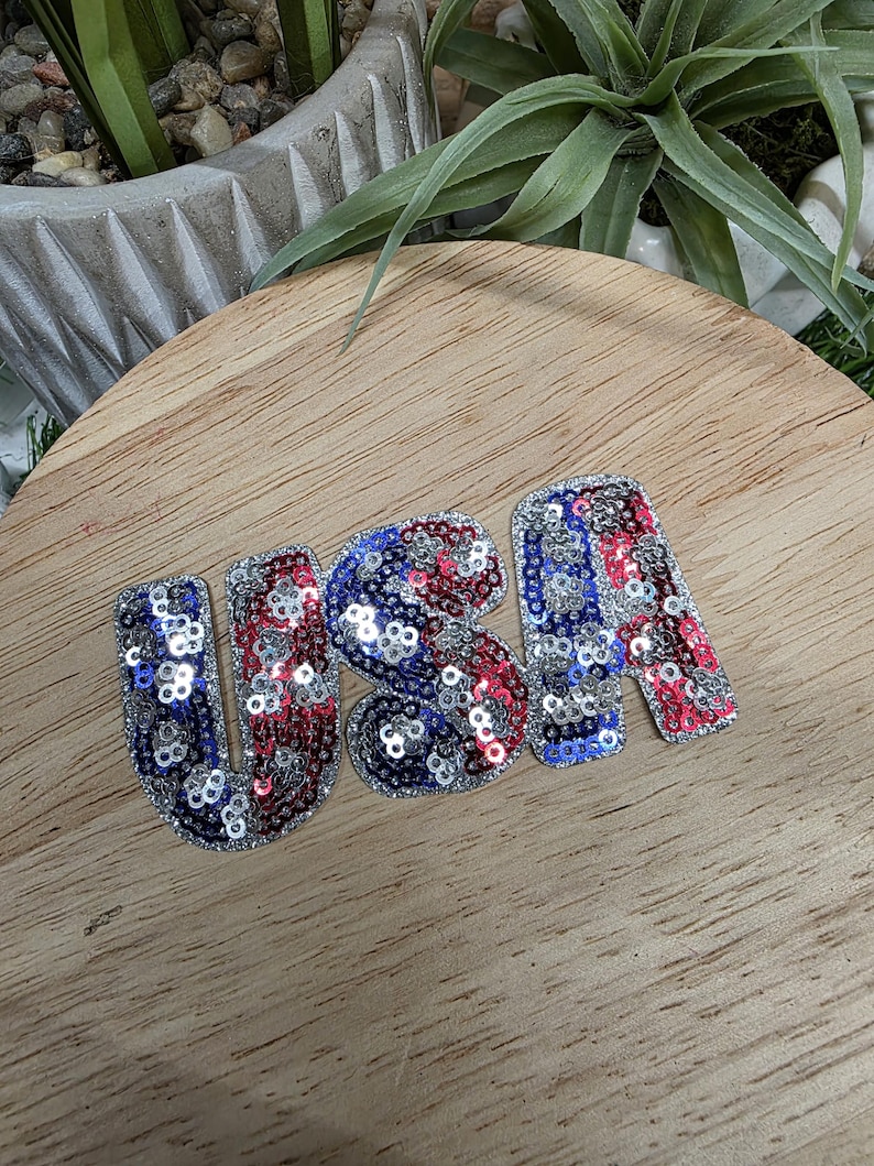 Pu&ograve; includere: Un emblema "USA" con paillettes rosse, bianche e blu, che ricorda la bandiera americana. Le lettere sono decorate con paillettes lucenti e sono poste su una superficie di legno. Si tratta di un oggetto decorativo patriottico.
