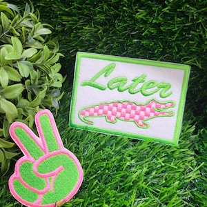May include: Two embroidered patches on a green background. One patch is a green and pink peace sign. The other patch is rectangular with the word "Later" in green script above a pink and white checkered alligator.