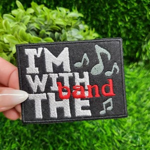 May include: Black rectangular patch with the text "I'M WITH THE band" in white and red lettering. The patch includes musical notes in a light green color. The patch is held against a background of green grass and foliage.