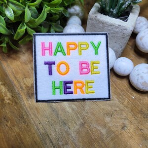 May include: A white embroidered patch with the text "HAPPY TO BE HERE" in rainbow colors. The patch is surrounded by a black border.