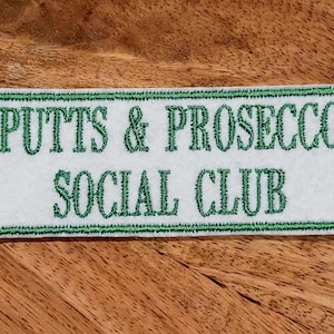 May include: White rectangular patch with green embroidered border. The text "PUTTS & PROSECCO SOCIAL CLUB" is embroidered in green.