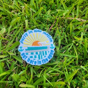 May include: A white embroidered patch with a blue boat, yellow sun, and the text "Party in slow motion out here in the open" with anchor symbols on either side.