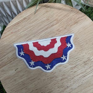 Pu&ograve; includere: Una toppa ricamata patriottica a forma di semicerchio. Il design presenta strisce rosse e bianche alternate, con un bordo blu ornato di stelle bianche. Per abbigliamento o accessori.