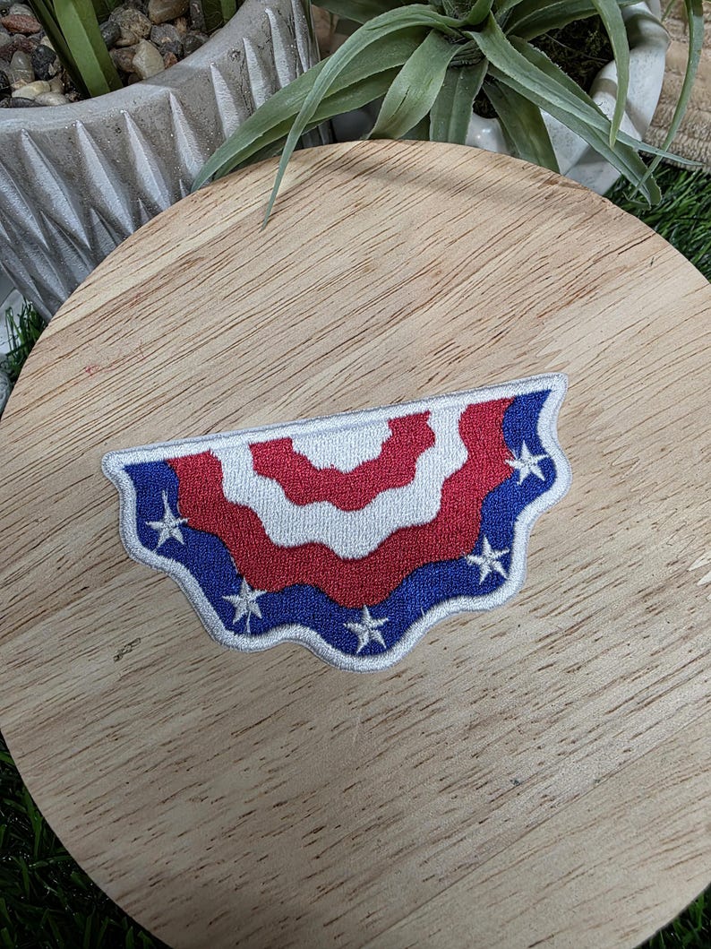 Pu&ograve; includere: Una toppa ricamata patriottica a forma di semicerchio. Il design presenta strisce rosse e bianche alternate, con un bordo blu ornato di stelle bianche. Per abbigliamento o accessori.