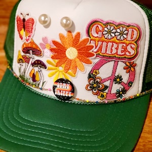 Puede incluir: Una gorra de camionero blanca con una visera verde. La gorra está decorada con parches coloridos, incluyendo un signo de paz, hongos, flores y las palabras "Good Vibes". También hay un botón que dice "Stay Trippy".