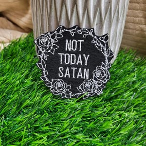 May include: Black embroidered patch with white stitching. The patch is shaped like a circle with a floral design around the edge. The text "NOT TODAY SATAN" is written in the center of the patch.
