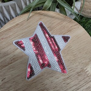 Pu&ograve; includere: Toppa a forma di stella con sfondo bianco e strisce diagonali di paillettes rosse. La toppa &egrave; su una superficie di legno. La stella ha un retro in feltro. Le paillettes catturano la luce, creando un effetto scintillante.