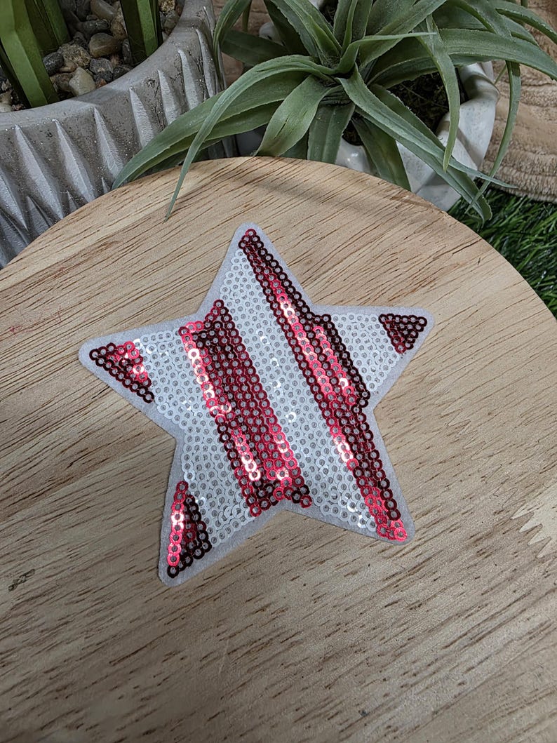 Pu&ograve; includere: Toppa a forma di stella con sfondo bianco e strisce diagonali di paillettes rosse. La toppa &egrave; su una superficie di legno. La stella ha un retro in feltro. Le paillettes catturano la luce, creando un effetto scintillante.