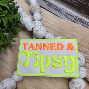 May include: A white patch with a neon green border and the text "Tanned & Tipsy" in orange and neon green lettering.