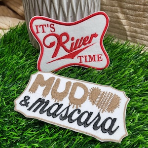 Puede incluir: Dos parches bordados. Uno es blanco con texto rojo que dice "It's River Time". El otro es blanco con texto marrón que dice "Mud & Mascara".