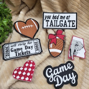 May include: A set of seven iron-on patches featuring football-themed designs. The patches include a red and white checkered heart, a brown football-shaped heart, a ticket stub with the text "Game Day Tickets", a black and white patch with the text "Game Day", a brown football with a red bow, a white elephant outline, and a black and white patch with the text "You had me at Tailgate".