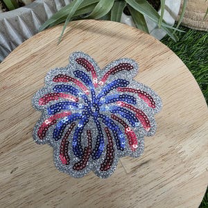 Pu&ograve; includere: Una toppa a forma di fuoco d'artificio con paillettes e un bordo argentato glitterato. Il design presenta paillettes rosse, bianche e blu disposte a raggiera, che ricordano un'esplosione di fuochi d'artificio.
