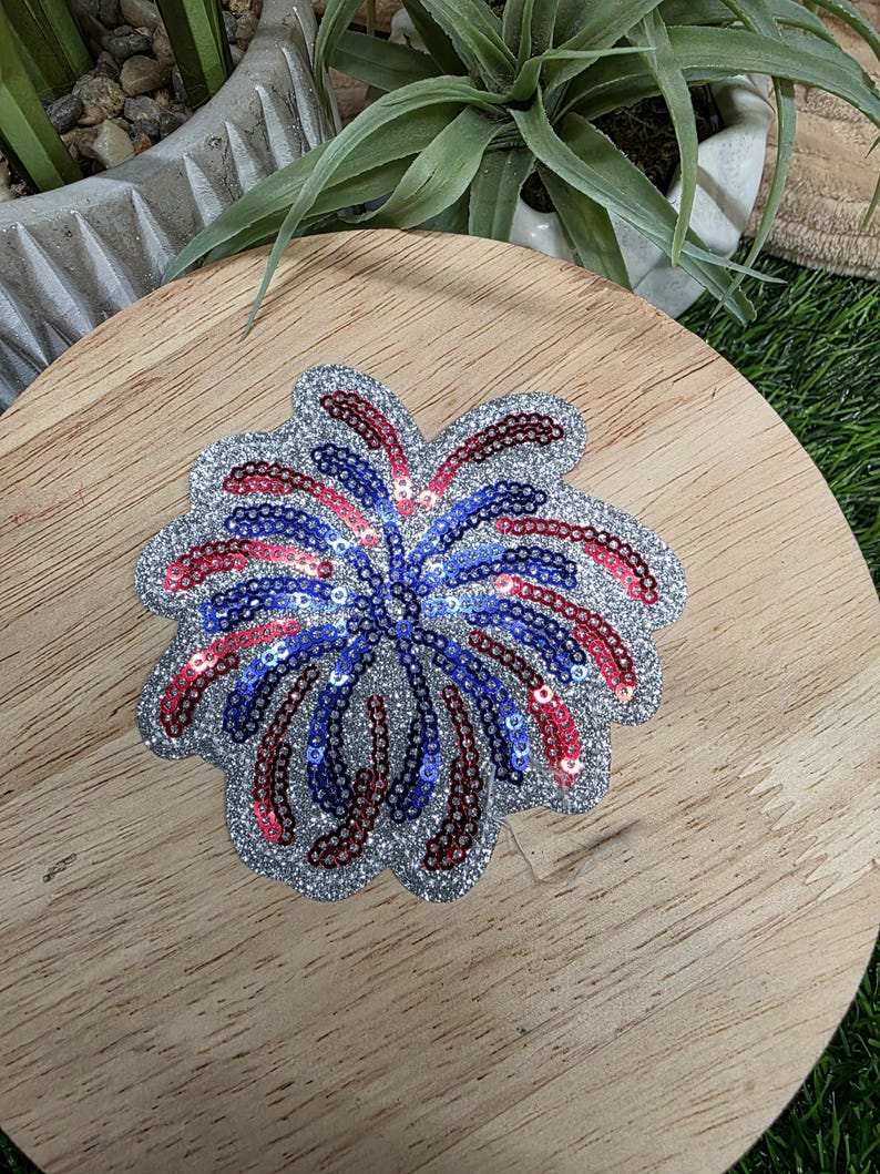 Pu&ograve; includere: Una toppa a forma di fuoco d'artificio con paillettes e un bordo argentato glitterato. Il design presenta paillettes rosse, bianche e blu disposte a raggiera, che ricordano un'esplosione di fuochi d'artificio.
