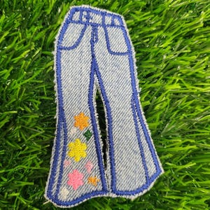 May include: A blue denim patch with a pair of bell bottom jeans embroidered on it. The jeans have a pocket and colorful flowers embroidered on the legs.
