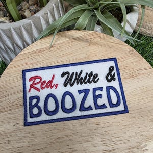 Pu&ograve; includere: Toppa ricamata con le parole "Red, White & BOOZED" in rosso, nero e blu. La toppa &egrave; rettangolare con sfondo bianco e bordo blu. La toppa &egrave; su una superficie di legno.