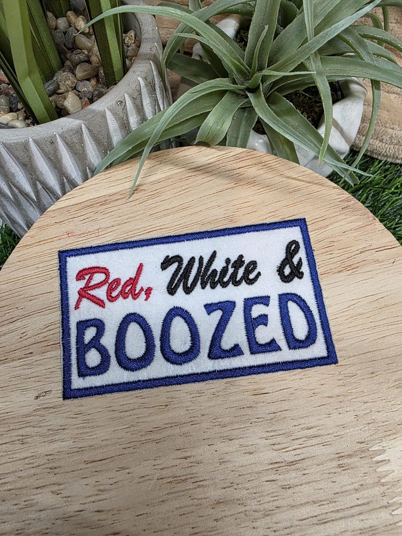 Pu&ograve; includere: Toppa ricamata con le parole "Red, White & BOOZED" in rosso, nero e blu. La toppa &egrave; rettangolare con sfondo bianco e bordo blu. La toppa &egrave; su una superficie di legno.