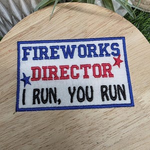 Pu&ograve; includere: Toppa ricamata con la scritta "FIREWORKS DIRECTOR I RUN, YOU RUN" in blu, rosso e nero. La toppa ha uno sfondo bianco e un bordo blu, con dettagli a stella. La toppa &egrave; su una superficie di legno.