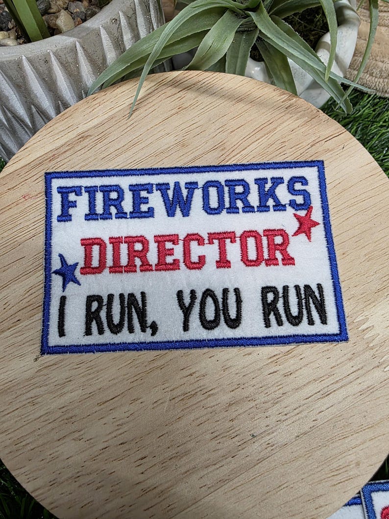 Pu&ograve; includere: Toppa ricamata con la scritta "FIREWORKS DIRECTOR I RUN, YOU RUN" in blu, rosso e nero. La toppa ha uno sfondo bianco e un bordo blu, con dettagli a stella. La toppa &egrave; su una superficie di legno.