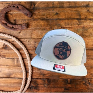May include: A grey and black flat bill snap back hat with a brown leather patch featuring a rooster wearing a cowboy hat. The patch has the text "RK" on the hat.