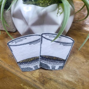 May include: Two embroidered patches of shot glasses with a black outline and a gold rim. The patches are on a wooden surface.