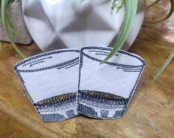 Op de afbeelding: Twee geborduurde patches van shotglazen met een zwarte omtrek en een gouden rand. De patches bevinden zich op een houten oppervlak.