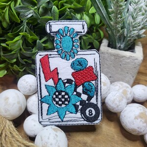 May include: Embroidered patch of a perfume bottle with a blue and white flower, red lightning bolt, black and white checkered pattern, and a blue and black eight ball.