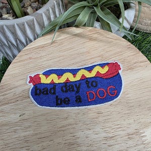 Pu&ograve; includere: Toppa ricamata a forma di hot dog, con panino blu, salsiccia rossa e senape gialla. Il testo "bad day to be a DOG" &egrave; ricamato in bianco e rosso.