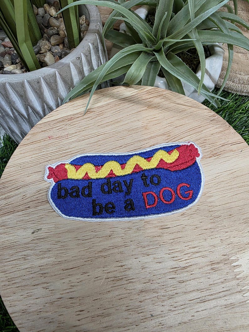 Pu&ograve; includere: Toppa ricamata a forma di hot dog, con panino blu, salsiccia rossa e senape gialla. Il testo "bad day to be a DOG" &egrave; ricamato in bianco e rosso.