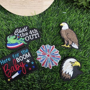 May include: A collection of embroidered patches with patriotic themes. Designs include a bald eagle, a firework, and phrases like "Chill the 4th OUT!" and "Here for the BOOM Baby!" on a green grass background.