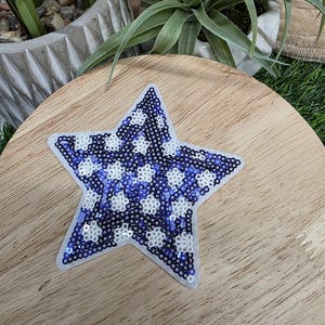 Pu&ograve; includere: Toppa a forma di stella con bordo bianco. La stella &egrave; riempita di paillettes blu e bianche, creando un motivo di forme floreali bianche su sfondo blu. La toppa &egrave; posizionata su una superficie di legno.