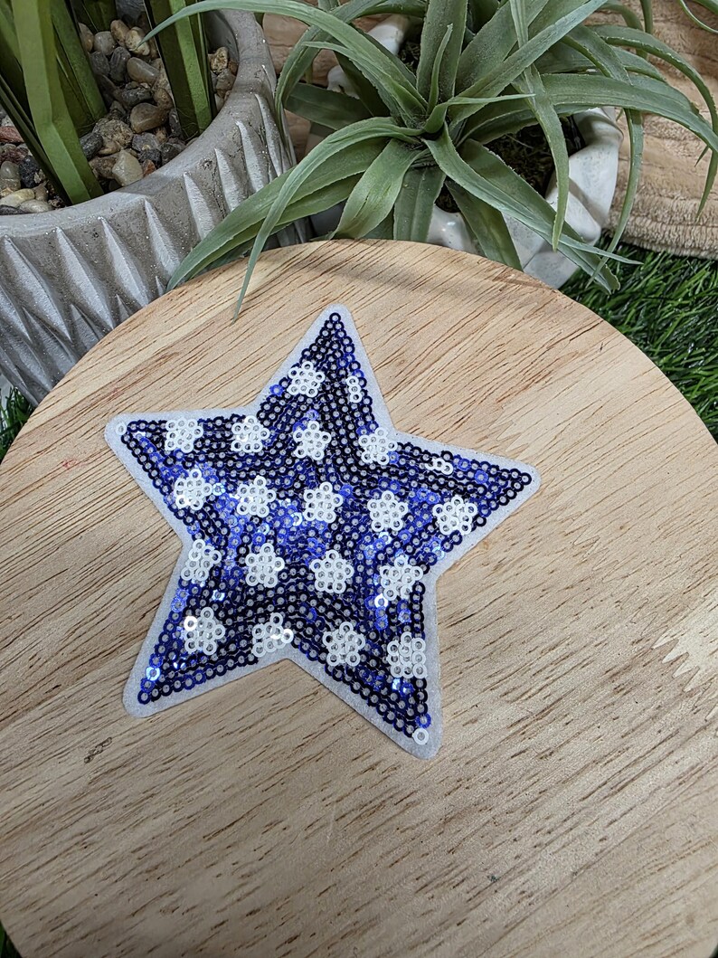 Pu&ograve; includere: Toppa a forma di stella con bordo bianco. La stella &egrave; riempita di paillettes blu e bianche, creando un motivo di forme floreali bianche su sfondo blu. La toppa &egrave; posizionata su una superficie di legno.