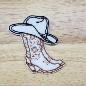 May include: Embroidered iron-on patch of a cowboy hat and boots. The hat is white with a black outline and the boots are brown with a floral design.