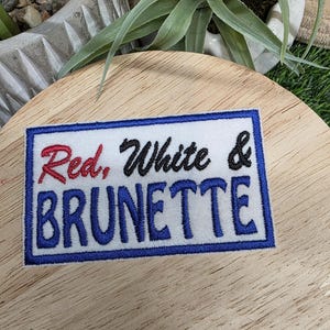 Pu&ograve; includere: Una toppa rettangolare bianca con bordo blu e le parole "Red, White & Brunette" ricamate in rosso, nero e blu. La toppa &egrave; su una superficie di legno.