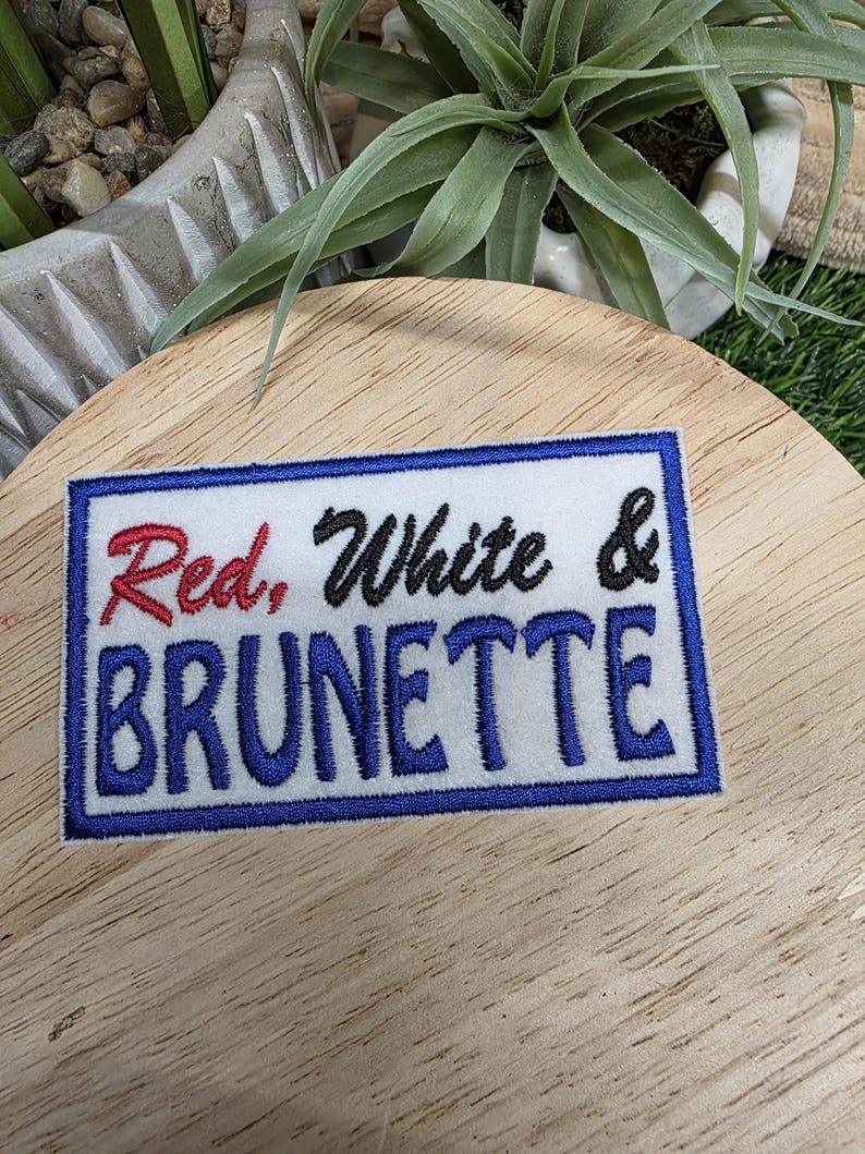 Pu&ograve; includere: Una toppa rettangolare bianca con bordo blu e le parole "Red, White & Brunette" ricamate in rosso, nero e blu. La toppa &egrave; su una superficie di legno.