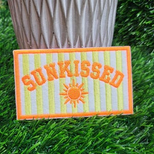 May include: Embroidered patch with the word "SUNKISSED" in orange, with a sun graphic, on a white and yellow striped background. The patch has an orange border and is set against a backdrop of green grass.