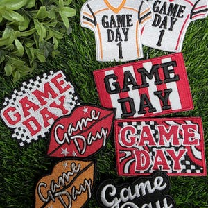 May include: A collection of embroidered patches featuring the text Game Day. Designs include two jersey-shaped patches, a red and white striped rectangle, a checkered pattern, two lip-shaped patches in red and orange, and a black script patch.