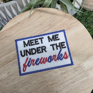 Pu&ograve; includere: Una toppa rettangolare con la scritta "MEET ME UNDER THE fireworks" in nero, rosso e blu. La toppa ha uno sfondo bianco con un bordo blu. La toppa &egrave; posizionata su una superficie di legno.