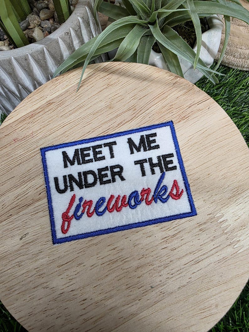 Pu&ograve; includere: Una toppa rettangolare con la scritta "MEET ME UNDER THE fireworks" in nero, rosso e blu. La toppa ha uno sfondo bianco con un bordo blu. La toppa &egrave; posizionata su una superficie di legno.