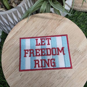 Pu&ograve; includere: Toppa ricamata con le parole "LET FREEDOM RING" in lettere rosse. La toppa ha uno sfondo bianco con strisce verticali blu e un bordo rosso. La toppa &egrave; su una superficie di legno.