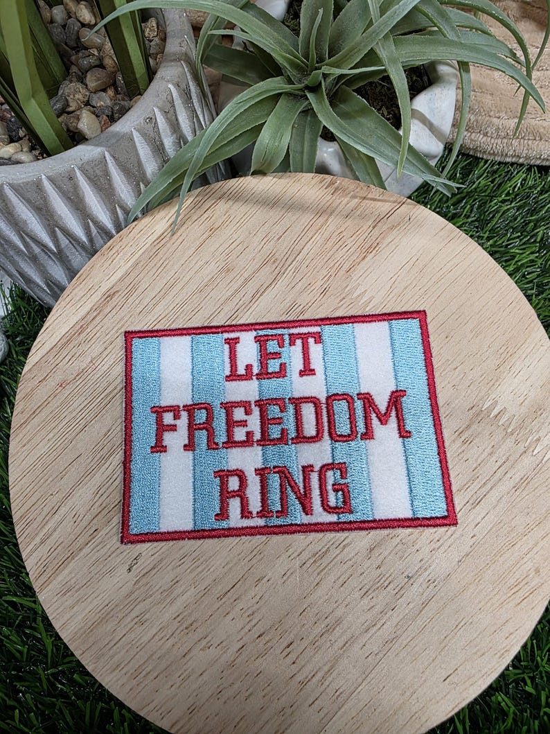 Pu&ograve; includere: Toppa ricamata con le parole "LET FREEDOM RING" in lettere rosse. La toppa ha uno sfondo bianco con strisce verticali blu e un bordo rosso. La toppa &egrave; su una superficie di legno.