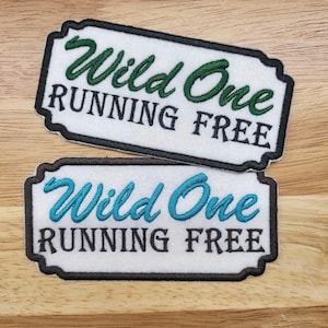May include: Two white fabric patches with black outlines. The top patch has green embroidery that reads "Wild One Running Free". The bottom patch has turquoise embroidery that reads "Wild One Running Free".
