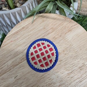 Pu&ograve; includere: Una toppa rotonda ricamata con bordo blu e motivo a scacchi rossi e bianchi. La toppa &egrave; centrata su una superficie di legno, con vegetazione sullo sfondo.