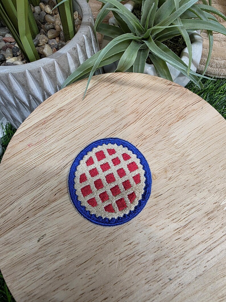 Pu&ograve; includere: Una toppa rotonda ricamata con bordo blu e motivo a scacchi rossi e bianchi. La toppa &egrave; centrata su una superficie di legno, con vegetazione sullo sfondo.