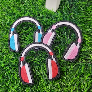 May include: Four embroidered headphone patches in blue, pink, and red with white and black accents. The patches are arranged on a bed of green artificial grass. The headphones have a curved top and ear pieces.