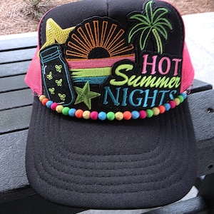 Könnte beinhalten: Schwarze Trucker-Mütze mit neonrosa Mesh-Rückseite und gebogenem Schirm. Die Vorderseite zeigt gestickte Aufnäher mit Sonnenuntergang, Palme und einem Glas mit Schmetterlingen. Der Text "HOT Summer NIGHTS" ist ebenfalls auf der Vorderseite gestickt.