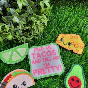 May include: A collection of embroidered food patches, including a lime wedge, a cheese slice, a taco, and an avocado with a smiling face. A patch with the text "FEED ME TACOS AND TELL ME I'M PRETTY!" is also included. The patches are on a green background.