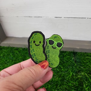 May include: Two embroidered green pickle patches. One pickle has a smiling face, and the other wears sunglasses. The patches are on a green, textured surface, with a white wooden background.