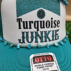Könnte beinhalten: Türkisblaue Trucker-Kappe mit einem weißen Frontpanel, auf dem der Text "Turquoise Junkie" in Schwarz steht. Die Kappe hat eine türkisblaue Schaumfront und ein Schweißband aus Frottee. Die Kappe hat einen goldenen Druckknopfverschluss und einen türkisblauen Perlenbesatz.