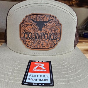 Pode incluir: Um boné de beisebol bege e marrom com um remendo de couro marrom com um design floral e o texto "COWPOKE". O boné tem uma aba plana e uma parte de trás de malha. O boné também tem uma etiqueta preta e vermelha com o texto "WEAR THE BEST", "RICHARDSON", "FLAT BILL SNAPBACK" e "EST 1970".